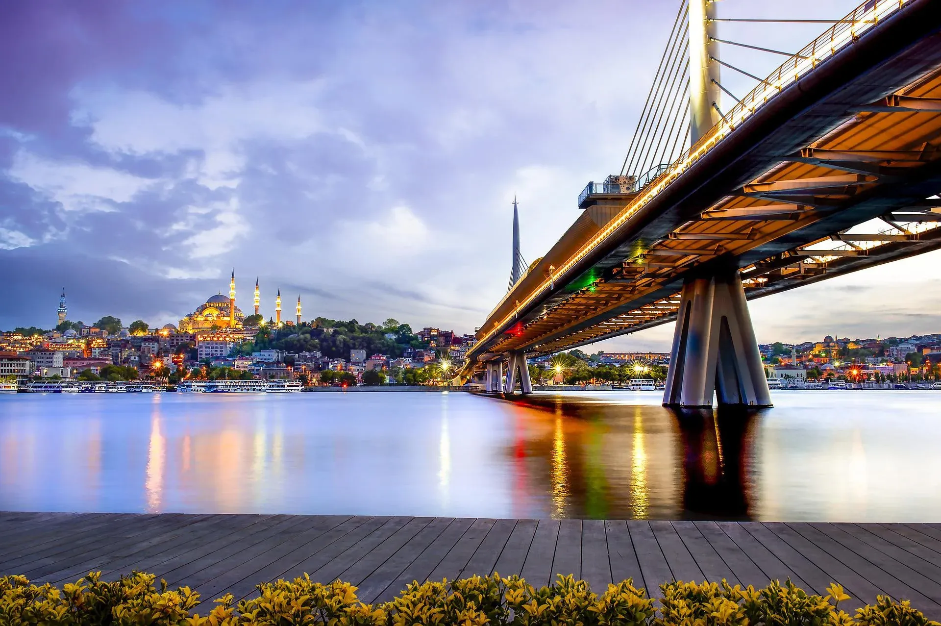 Illuminated Golden Horn Metro Bridge over water at dusk, with the Süleymaniye Mosque and Istanbul cityscape in the background