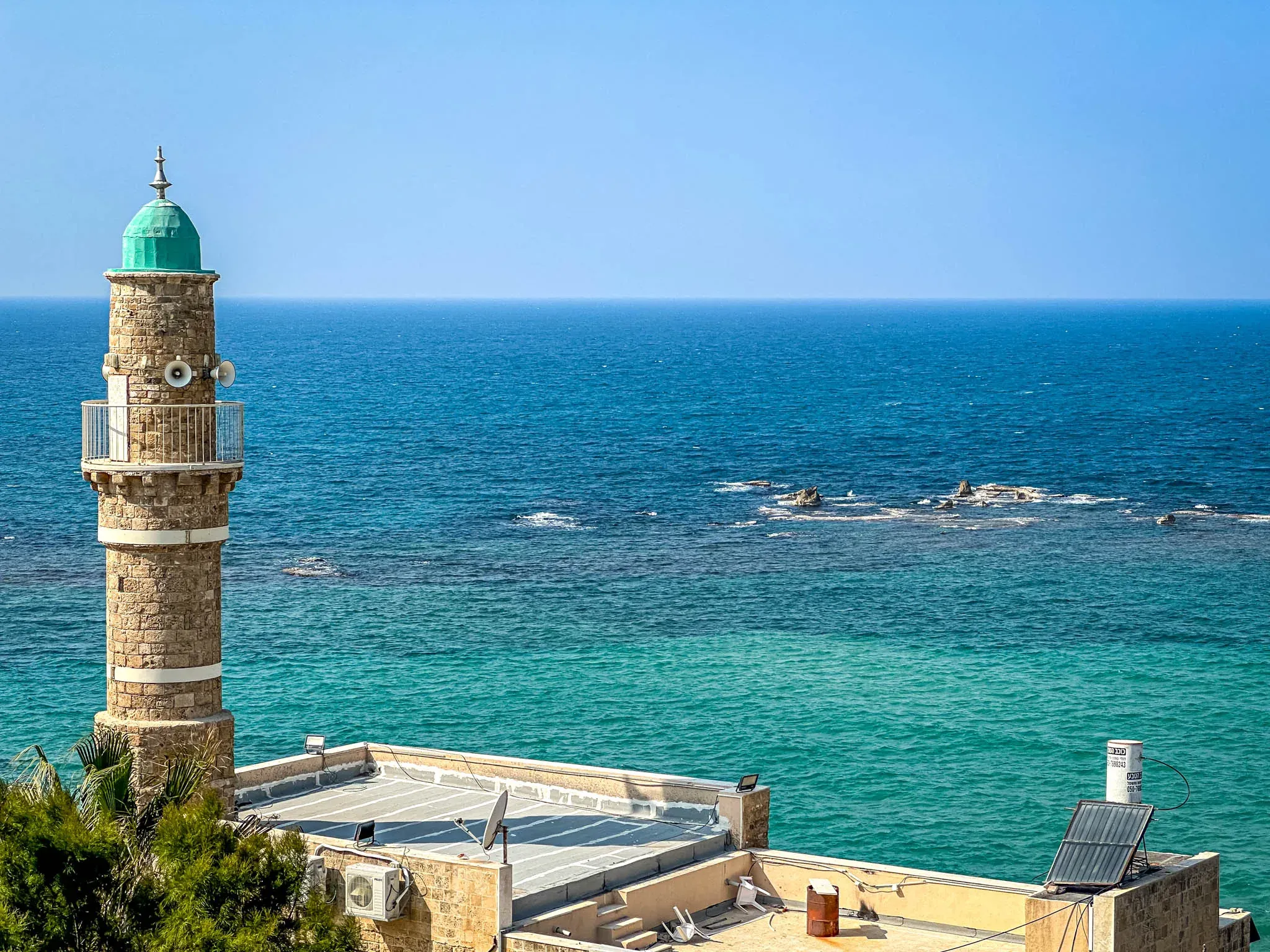 Stone minaret with a green dome and balcony overlooking the turquoise Mediterranean Sea, with rooftops and green foliage.