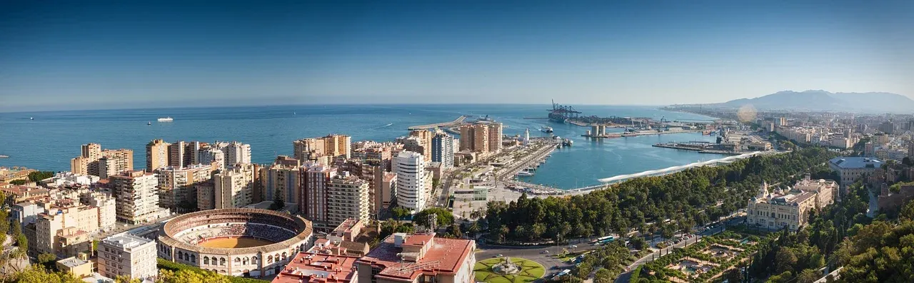 Panoramic view of Malaga cityscape with La Malagueta bullring, the port, and the Mediterranean Sea under a blue sky.
