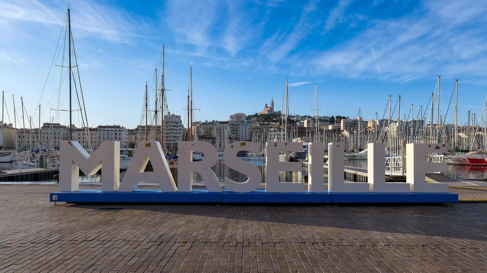 Large MARSEILLE letters on a blue base at the Vieux-Port, with sailboats and buildings. Notre-Dame de la Garde visible on the