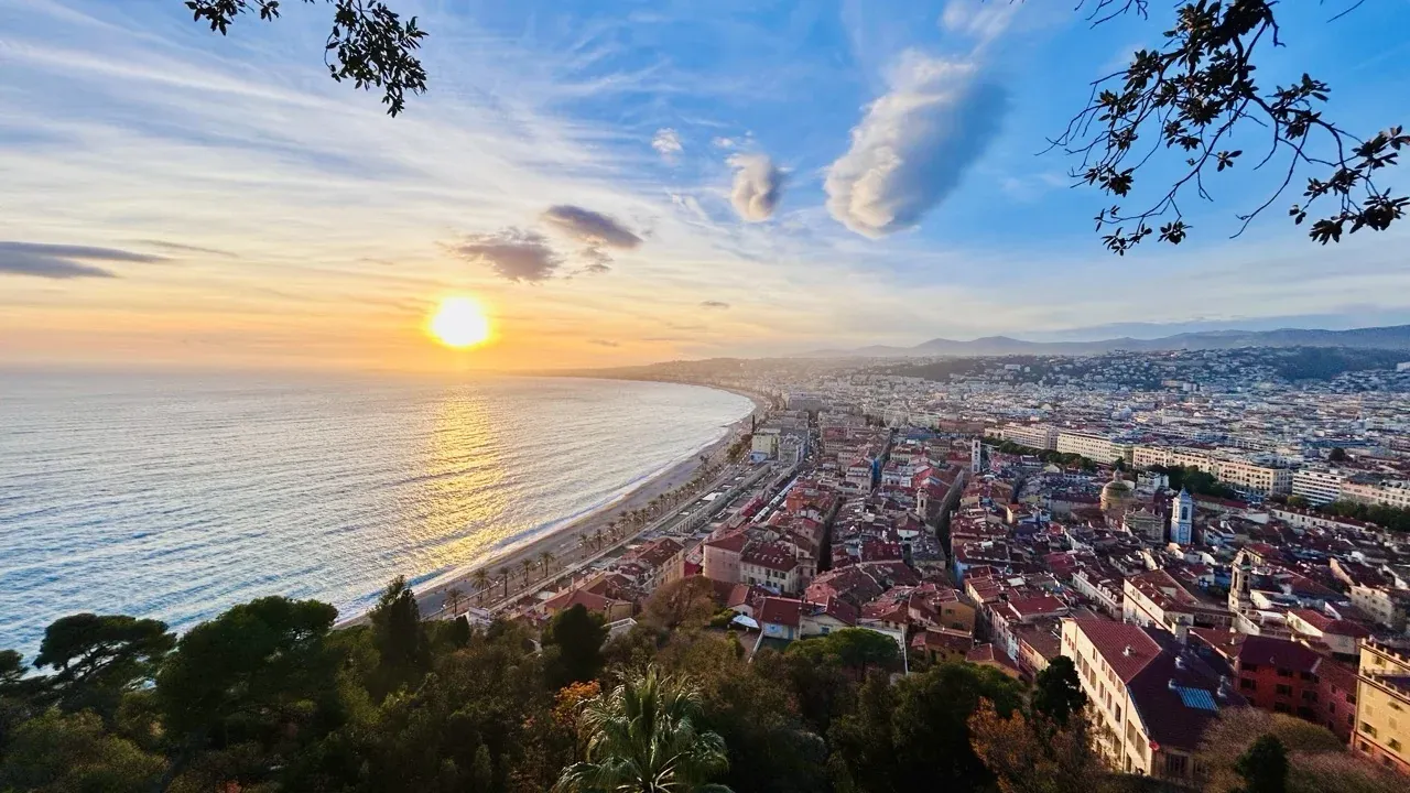 Panoramic view of Nice coastline at sunset, showing the sun reflecting on the sea, a long beach, promenade, and the city buil