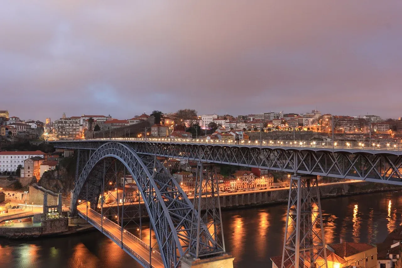 Large metal arch bridge with two decks illuminated at dusk over a river, with city buildings on both sides.