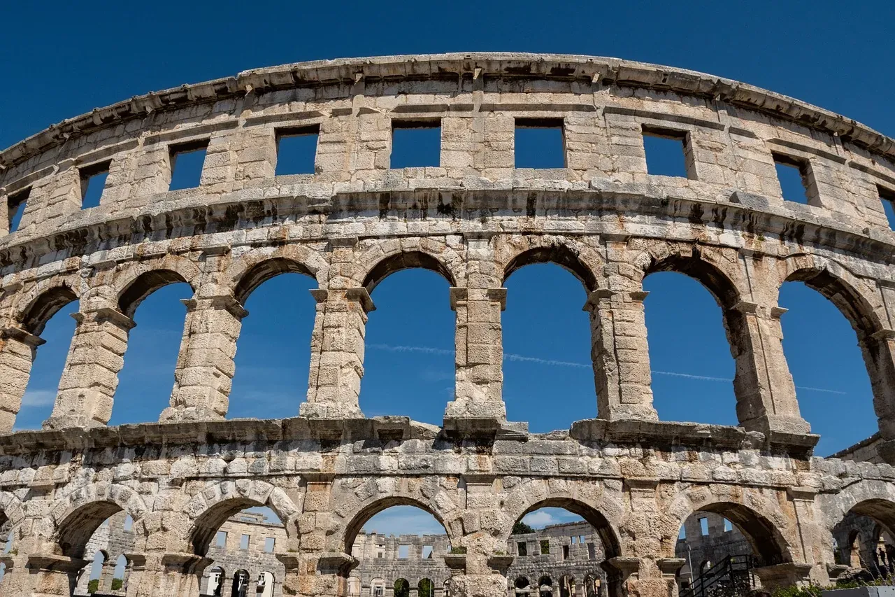 Exterior view of the Pula Arena, an ancient Roman amphitheater with multiple arched levels and windows against a clear blue s