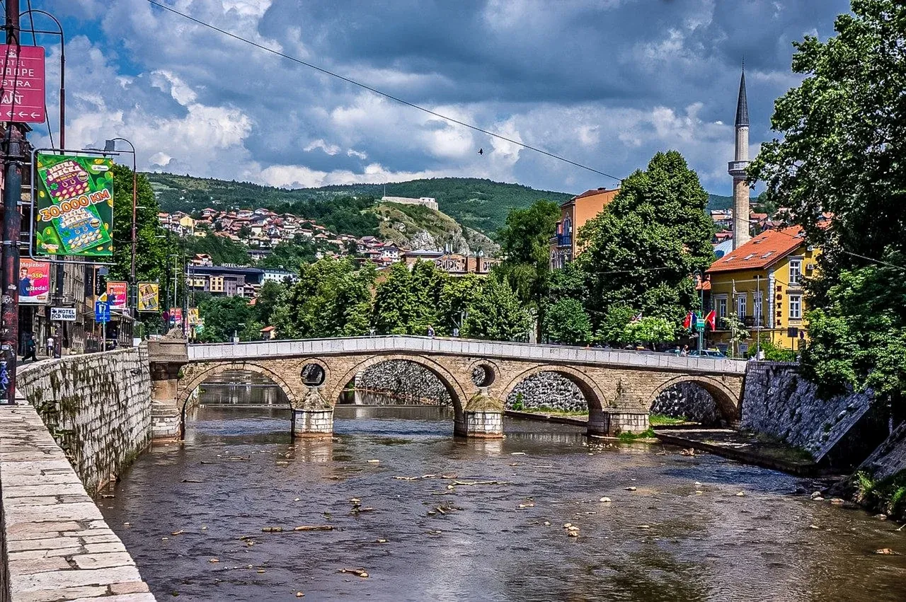 Historic stone arch bridge over a river in Sarajevo, with a minaret, buildings, and green hills under a cloudy sky.