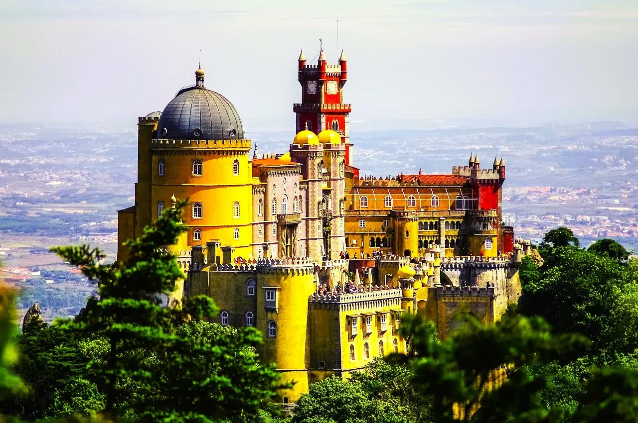 Colorful Pena Palace in Sintra, Portugal, featuring yellow, red, and grey sections with domes and towers, seen from afar thro