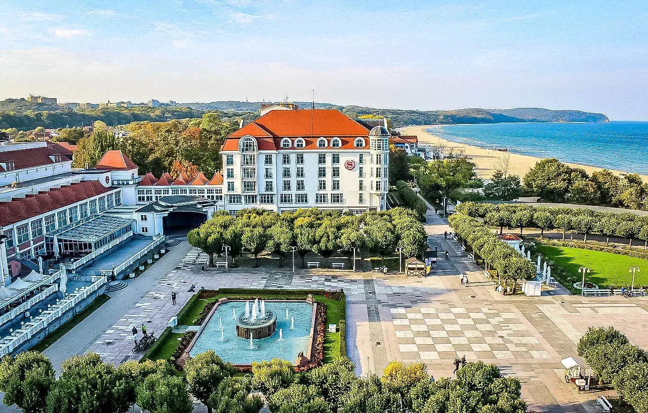 Aerial view of Grand Hotel Sopot with a square, fountain, green trees, sandy beach, and Baltic Sea under a blue sky.