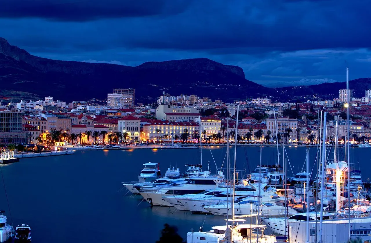 Harbor at twilight with numerous yachts and boats, illuminated waterfront buildings, palm trees, and mountains under a dark b
