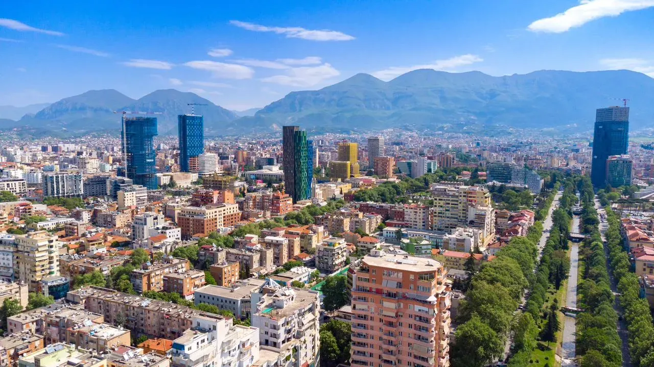 Aerial view of Tirana city with modern high-rise buildings, residential areas, a tree-lined boulevard, and mountains.