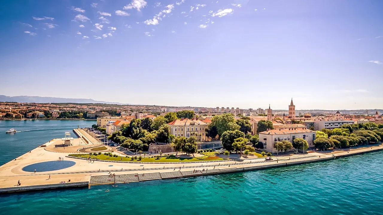 Aerial view of Zadar waterfront with the Sea Organ, old town buildings, clear blue sea, and distant mountains under a sunny s
