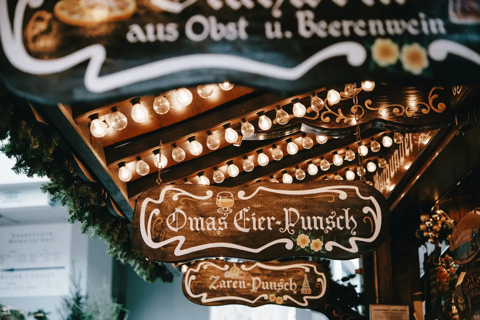 Wooden Christmas market signs advertising Eierpunsch and other drinks decorated with strings of warm lights in Berlin.