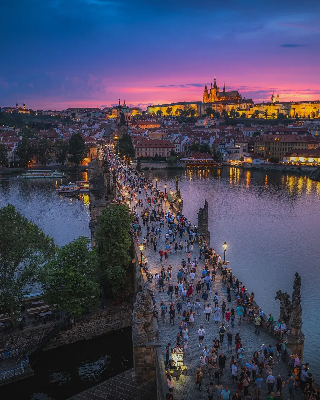 Tourists stroll across Charles Bridge in Prague at sunset, with Prague Castle majestically lit in the background.