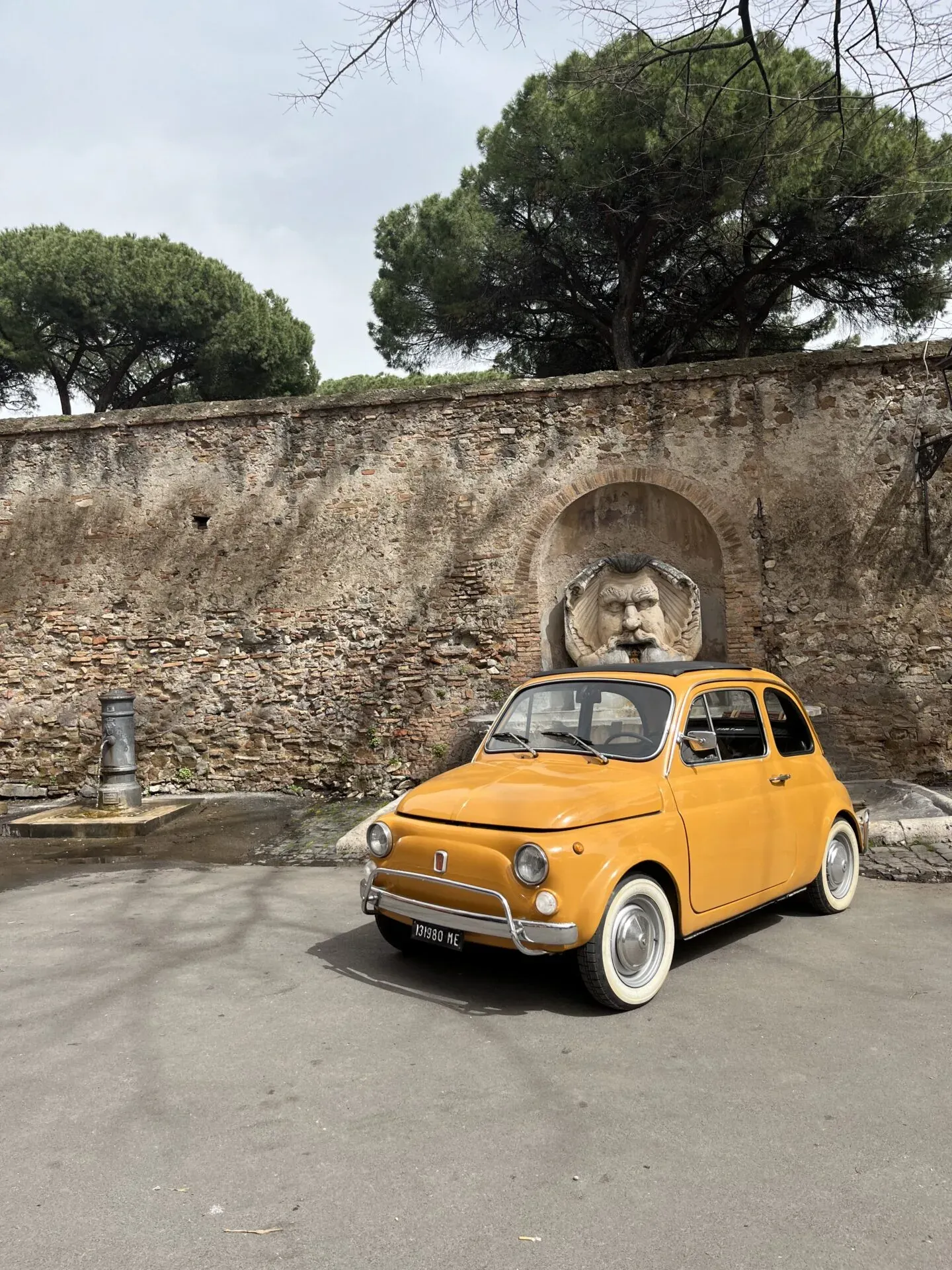A yellow Fiat 500 parked near a Roman wall with a unique fountain.